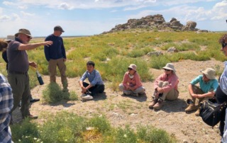 Archaeologist giving an outdoor lecture at Pınarbaşı, with students listening and the surrounding rocky landscape in the background.
