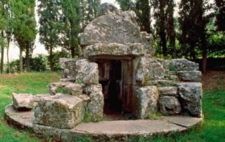 La Tanella di Pitagora, an ancient Etruscan tomb in Cortona, Italy, surrounded by lush greenery and featuring a circular stone structure with an entrance to the burial chamber.
