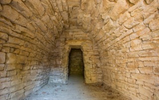 Interior of Tumulus 2 at the Area Archeologica del Sodo in Cortona, Italy, showcasing an ancient Etruscan burial chamber with stacked stone walls and a passageway leading to the main chamber.