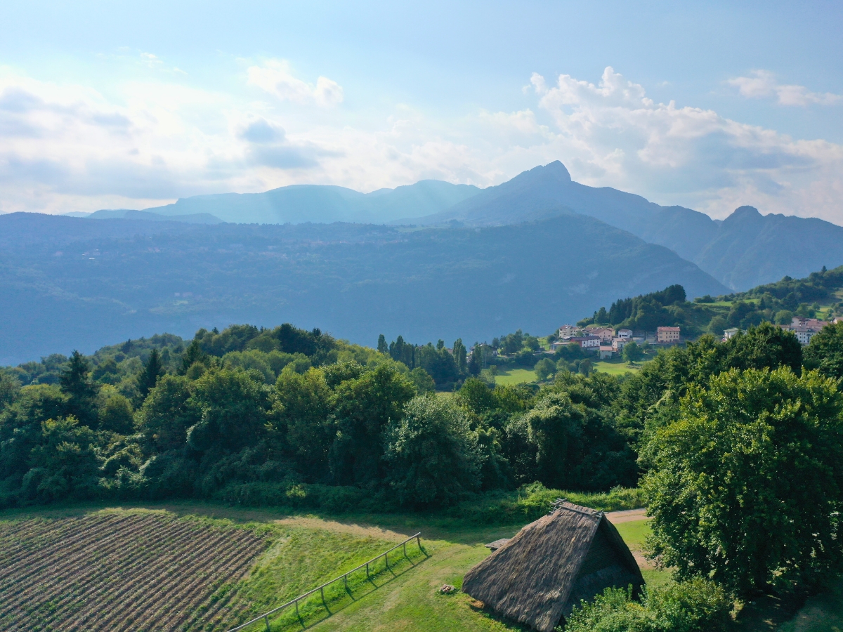 Aerial view of a traditional thatched hut and surrounding landscape at Bostel di Rotzo, with lush green fields, forest, and mountains in Vicenza, Italy.