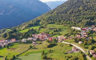 Aerial view of Bostel di Rotzo in Vicenza, Italy, showing a scenic village nestled in green fields and surrounded by forested mountains.