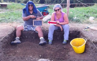 Archaeology students and instructors at a field excavation site in Bostel di Rotzo, Italy, holding an excavation sign and equipment.