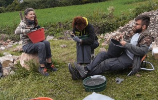 Archaeology students sifting soil and examining finds at Bostel di Rotzo, Italy, while seated on stones.