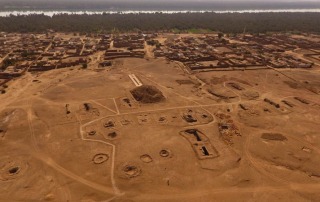 Aerial drone shot of the El Kurru archaeological site in Sudan, showing an expansive view of ancient ruins, excavation areas, and surrounding village landscape