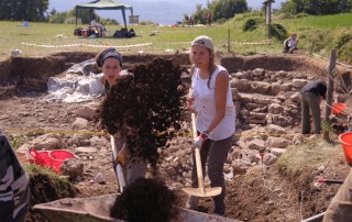 Students at the Bostel di Rotzo field school in Italy, excavating a site and gathering archaeological materials, with a backdrop of rolling hills.
