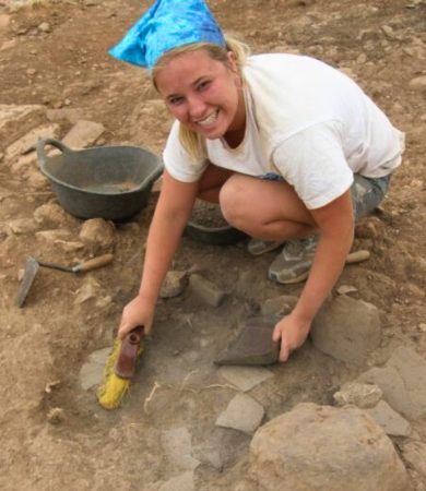 A young Kate is excavating during her first field school in Menorca, Spain. She is smiling while using tools to uncover artifacts.