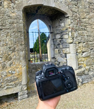 Close-up of a digital camera held up to photograph a gable at an archaeological site, used for photogrammetry by a student documenting archaeological details