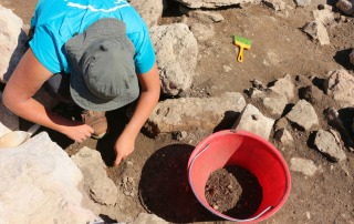 Overhead view of a student working at an archaeological excavation site with tools and a red bucket, capturing field school activities in Vicenza, Italy.