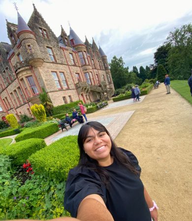 Smiling student taking a selfie in front of Belfast Castle, with stunning architecture and well-manicured gardens, during a field trip in Northern Ireland