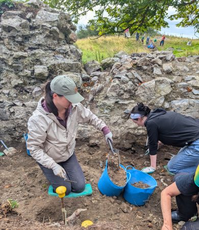 Students participating in an archaeology field school in Ireland, excavating historical ruins with tools and blue buckets