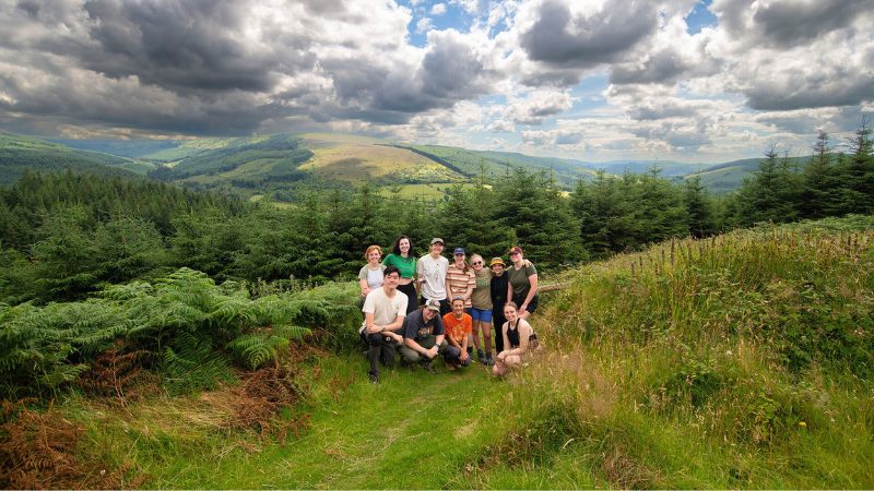 A group of archaeology field school students posing together in a scenic, mountainous landscape in Ireland, surrounded by lush greenery and under a dramatic cloudy sky