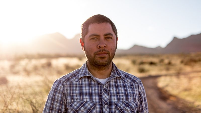 Jason De Leon standing in a Chiapas field at sunset, wearing a plaid shirt, with mountains in the background.