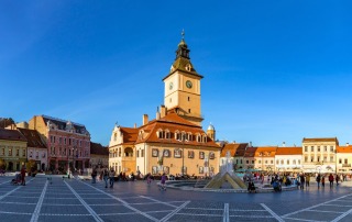 Panoramic view of the Brasov Town Square in Romania, featuring the iconic Clock Tower, historic buildings, and a bustling community atmosphere under a clear blue sky.