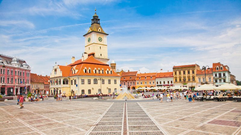 A vibrant view of the Brasov Town Square in Romania, featuring historic architecture and a fountain, a must-visit location during the Romania Sanpetru Program.