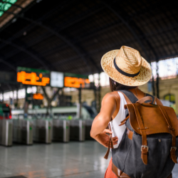 A traveler with a hat and backpack looking at train schedules in a station, emphasizing travel readiness.