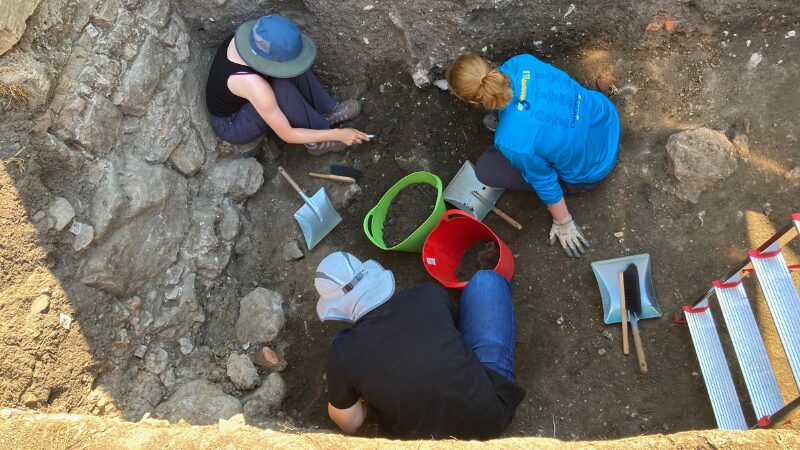 Students excavating at archaeological pit at Sanpetru field school in Romania Overhead view of three students working together in an archaeological excavation pit at the Sanpetru site in Romania, with various tools and buckets surrounding them.