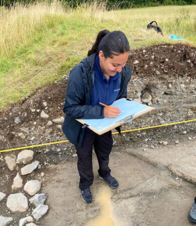 Taking elevations at Clonoghill Castle An archaeology student taking elevation measurements at the Clonoghill Castle excavation site, using a clipboard and standing beside a partially excavated trench.