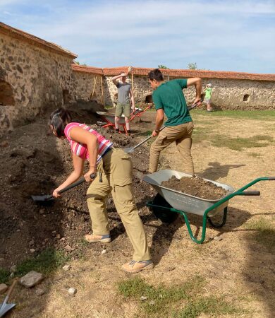 Students moving excavated earth at Sanpetru, Romania Two students using shovels and a wheelbarrow to move excavated soil at the Sanpetru archaeological site, with stone walls of the dig area in the background.
