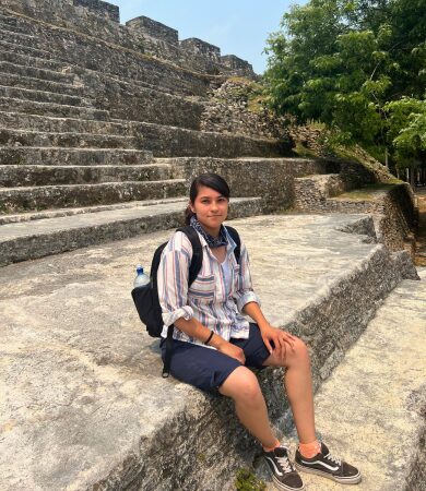 Archaeology student sitting on ancient Maya ruins at an excavation site in Belize, wearing a striped shirt and backpack.