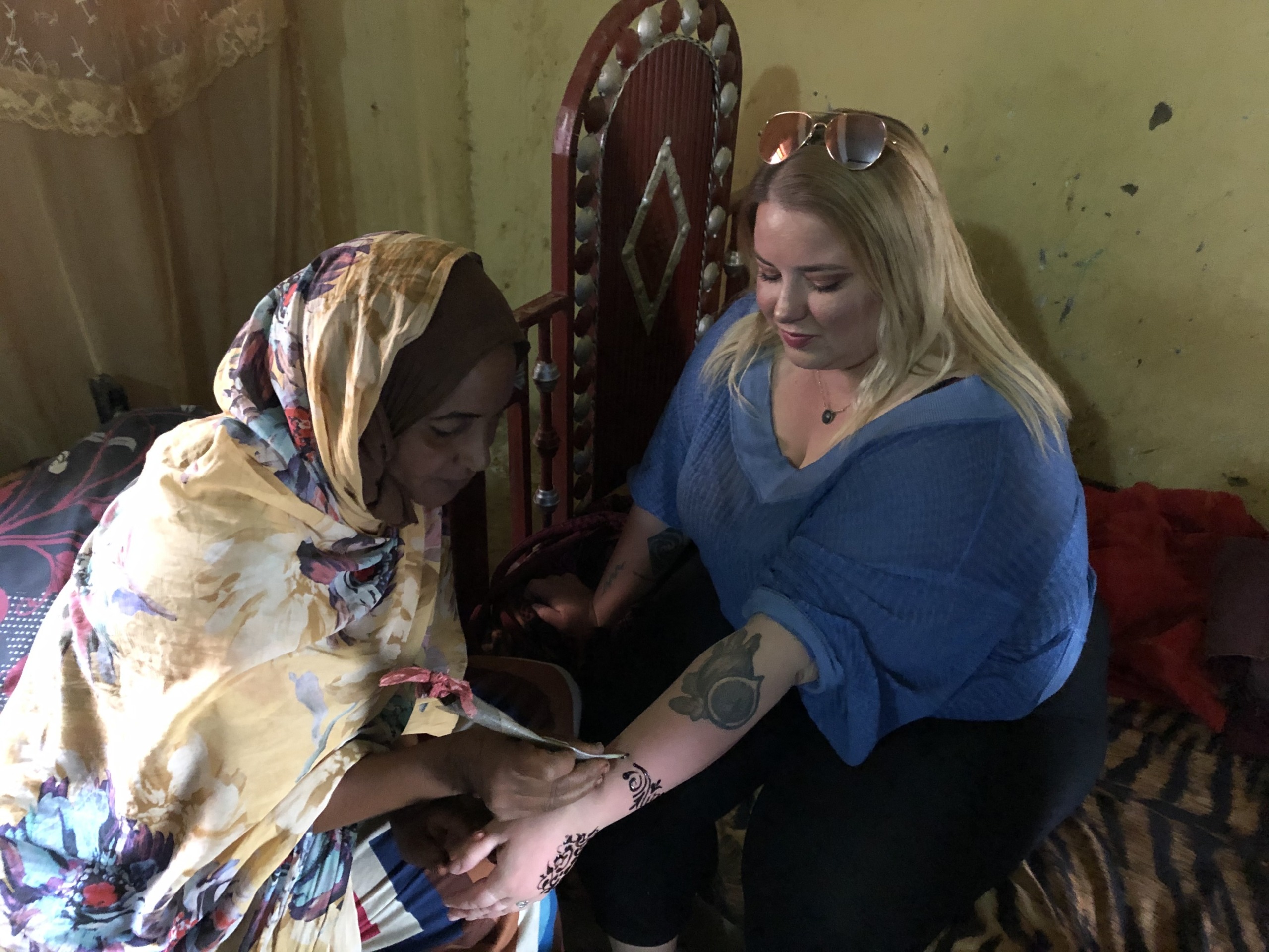 A woman in a blue top sits while another woman in a floral headscarf applies traditional henna designs to her arm inside a warmly lit room in Sudan.
